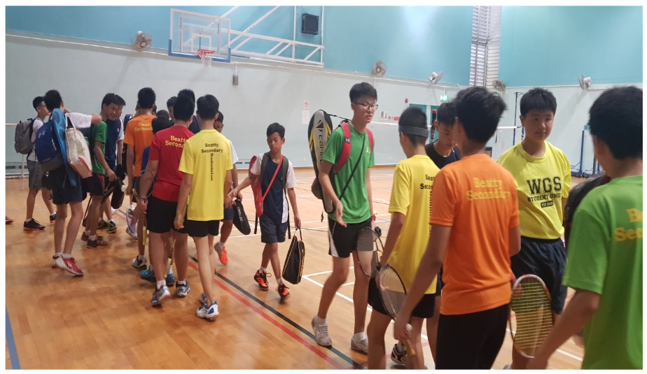 Students in colorful jerseys walk across an indoor badminton court carrying rackets and bags.